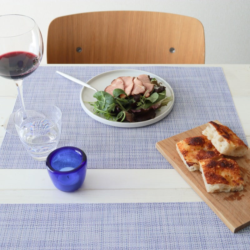 「Chilewich Mini Basketweave ランチョンマット(ペリウィンクル)」の上に白いプレートとカトラリー、赤ワイン、ブルーのキャンドルが配置されたモダンなテーブルスタイリング。