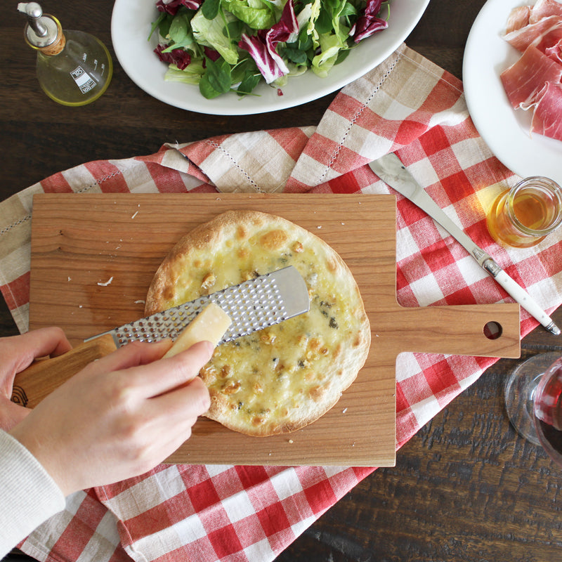 ブラックチェリー材を使用したダスホルツのシンプルな カッティングボードスクエア でトルティーヤピザを作って仕上げにチーズを削っている。サタルニアの食器やビアンキのグレーターを使ってレストランの様な演出。